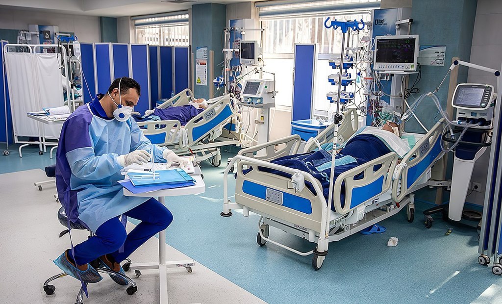 Two Coronavirus patients hooked up to ventilators. A doctor in PPE is writing charts at a small rolling table in the foreground.