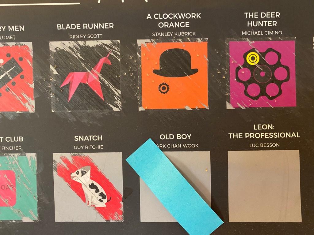 6 squares on the 100 Movies Busket List poster. 3 are all the way scratched off: Blade Runner, A Clockwork Orange, The Deer Hunter. 1 is halfway scratched off: Snatch. 2 are untouched: Old Boy and Leon: The Professional. The square for Old Boy is marked with a post-it flag.
