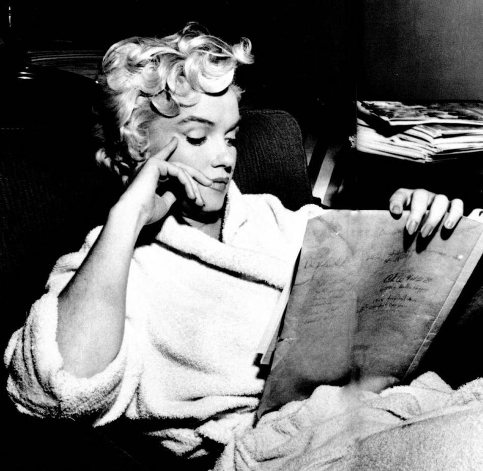 A black and white photo of Marilyn Monroe, in a white terrycloth bathrobe. She has a pensive look on her face as she reads, looking at the book in her hand rather than at the camera.