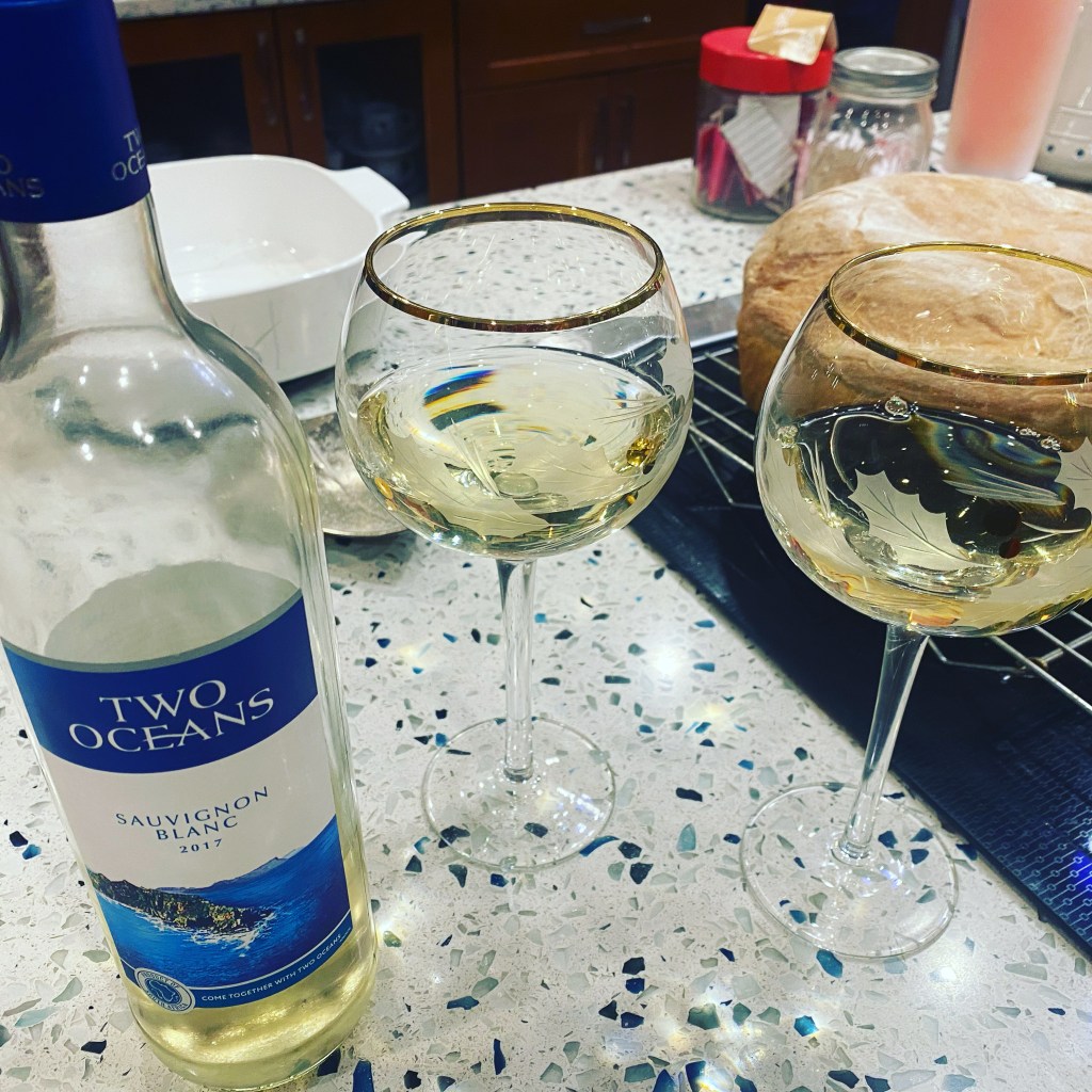 A bottle of white wine next to two filled wine glasses. In the background, a fresh-baked loaf of bread sits on a cooling rack.
