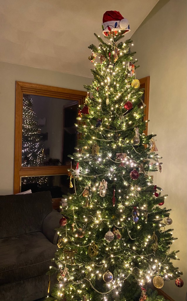 A Christmas tree with white lights and decorations, topped with a Santa Claus hat. The tree and white lights can also be seen reflected in a window behind it.