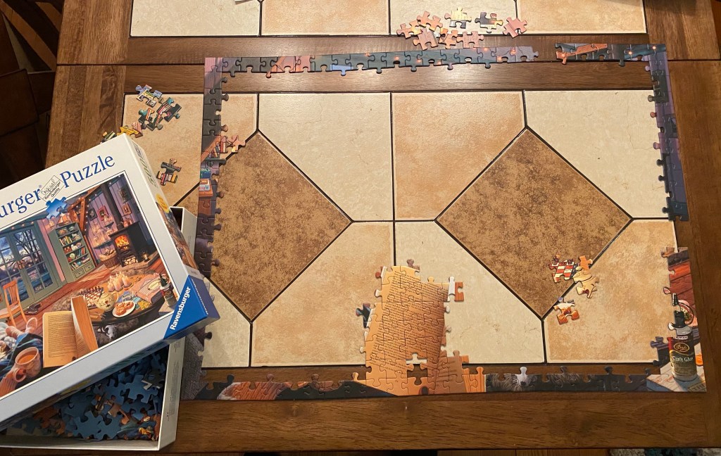 Picture of a jigsaw puzzle being assembled. The edge is mostly done, and the box of puzzle pieces is to the left of the puzzle-in-progress.