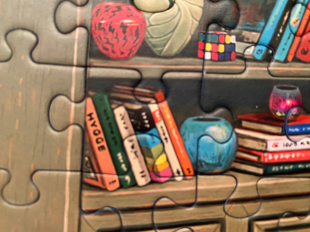 Close-up of a completed jigsaw puzzle, showing bookshelves holding vases, a rubik's cube and several books--the one book with a readable title says "Hygge"