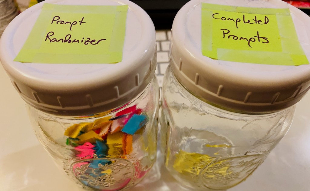 Two mason jars sitting side by side. The one on the left is labelled "Prompt Randomizer" and contains many small bits of folded paper. The one on the right is labelled "Completed Prompts" and has one slip of paper in it.